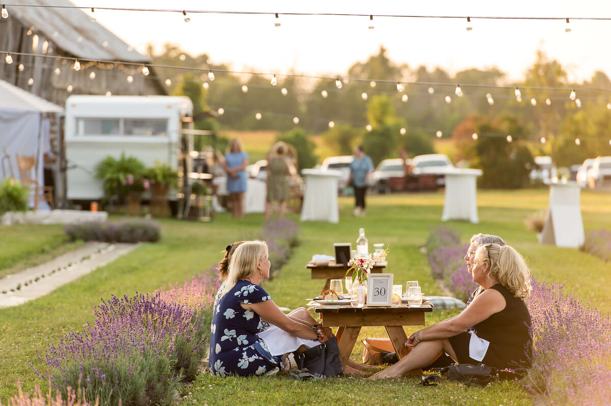 a photo of women sitting and enjoying charcuterie with the bar and twinkle lights in the background as part of Soiree in the Field.  Captured by Ottawa Event Photographer JEMMAN Photography COMMERCIAL