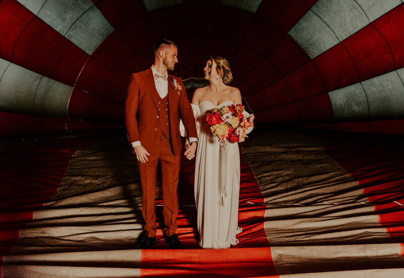 Mariés de plein pied et de face, le regard tourné l'un vers l'autre à l'intérieur d'un ballon de montgolfière bariolé rouge et blanc. Par Laura Termeau photographie.