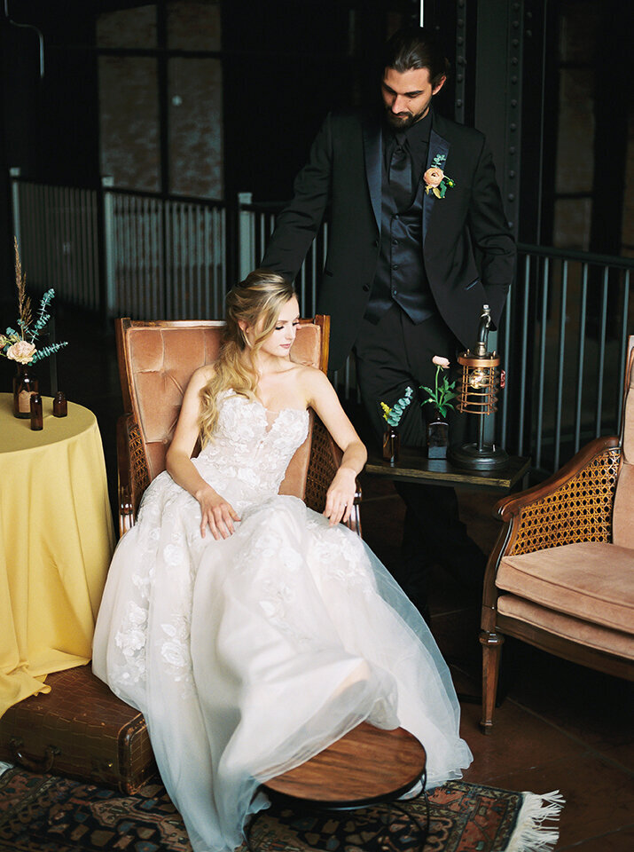 Bride wearing a white wedding gown sitting on a blush-colored chair with groom standing behind her wearing a black tuxedo.