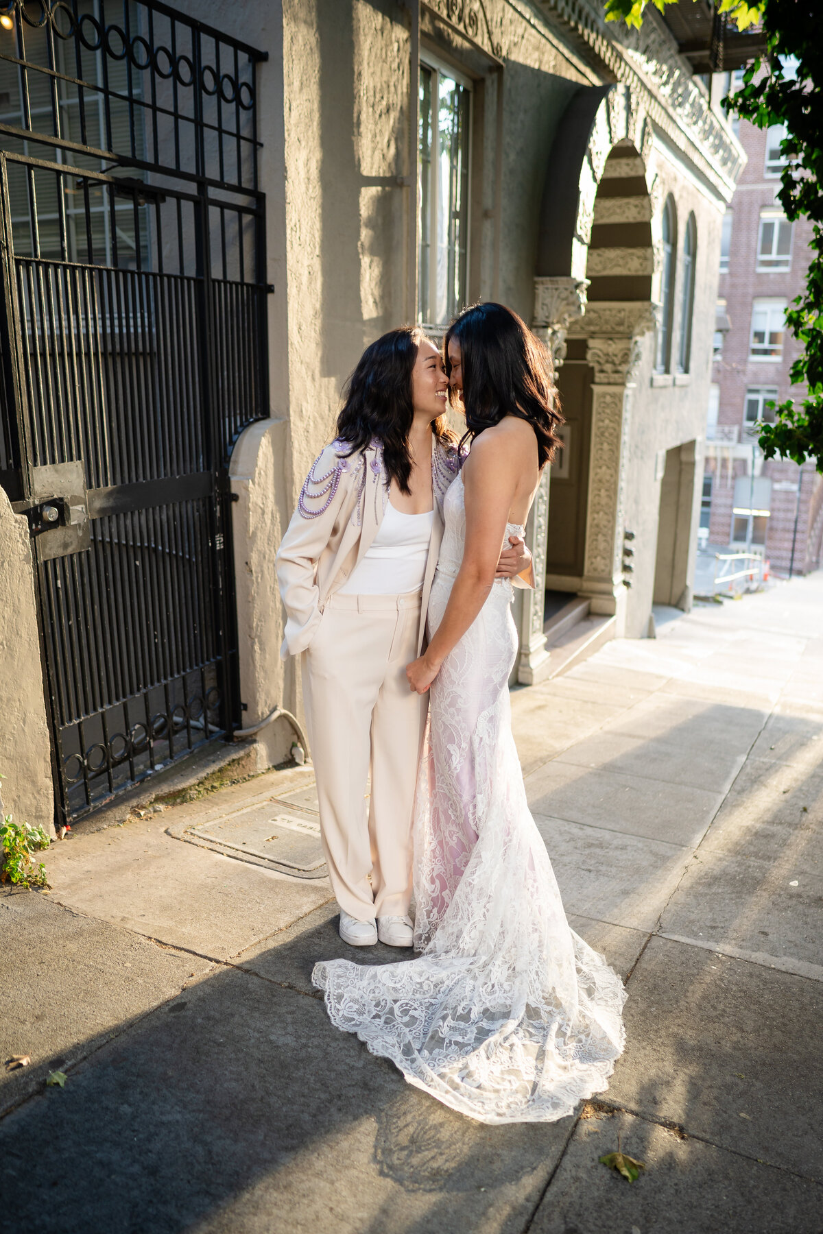 Queer lesbian brides in San Francisco during sunrise on the street giving each other eskimo kisses in their purple accent outfits, one wearing the Quill gown by Claire Pettibone