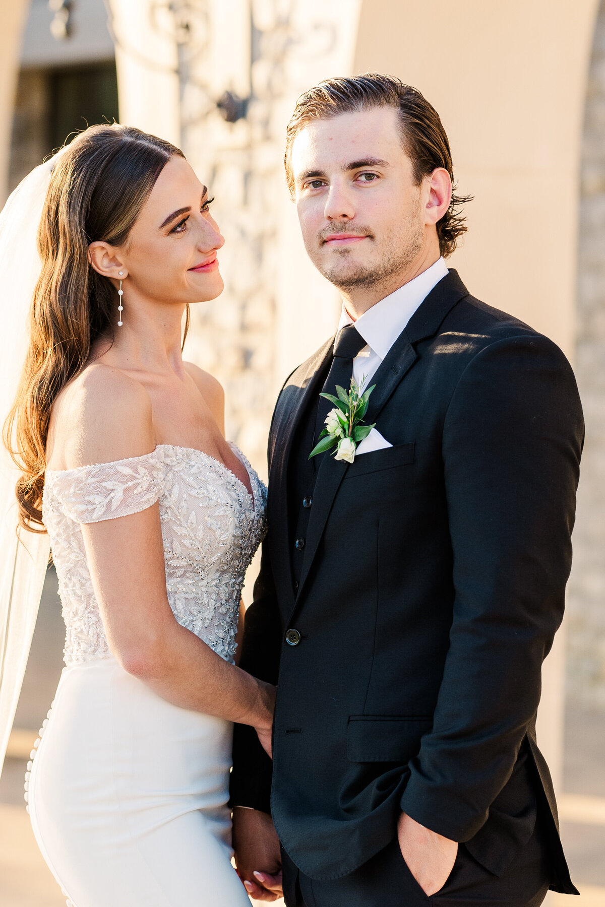 Elegant bride in a beaded off-the-shoulder gown stands beside her groom in a black tuxedo, softly lit by golden hour light outside a stone estate in Westlake, Texas.