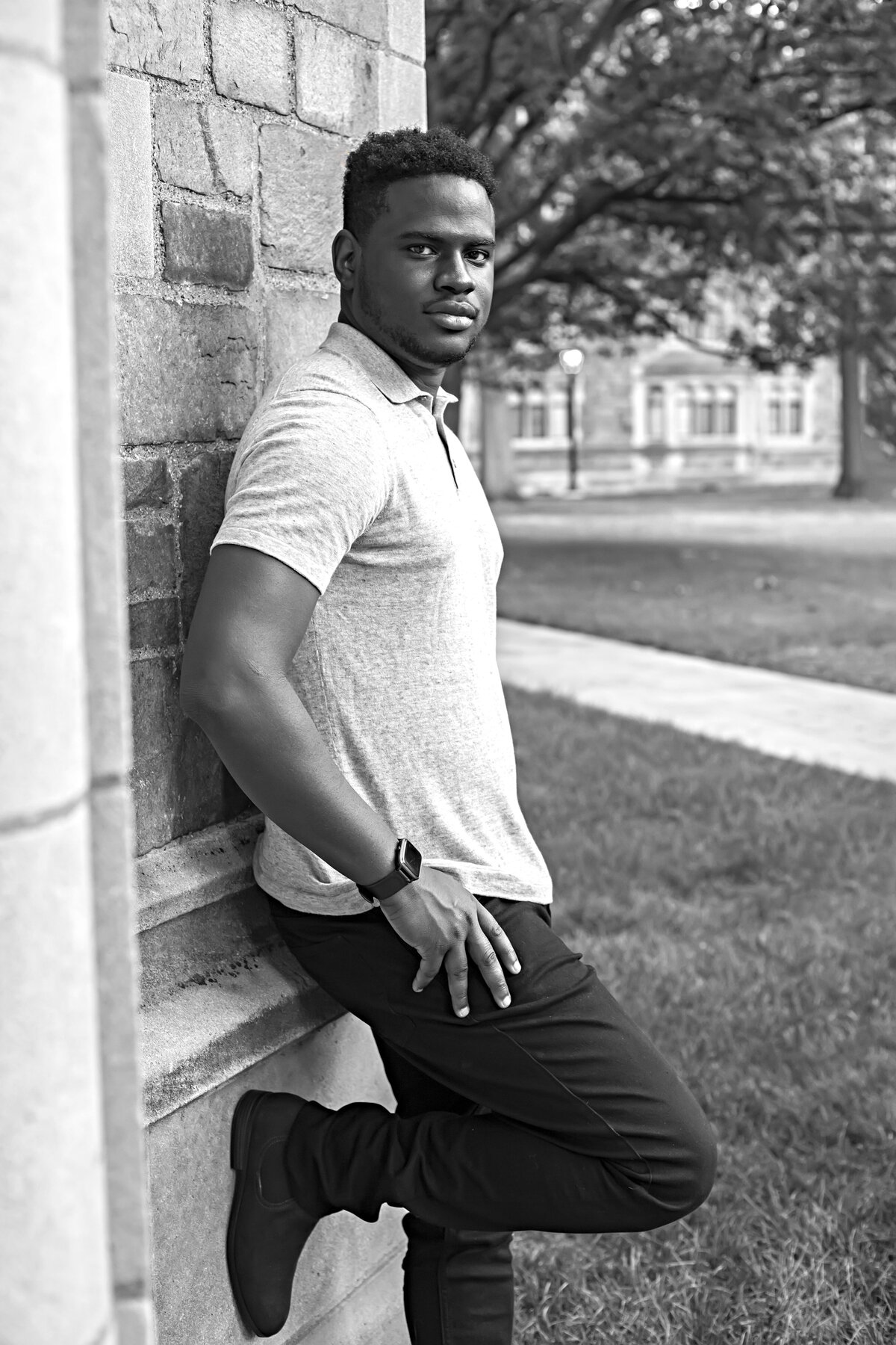African American male wearing tan polo shirt, black jeans, and black shoes leaning against a brick wall
