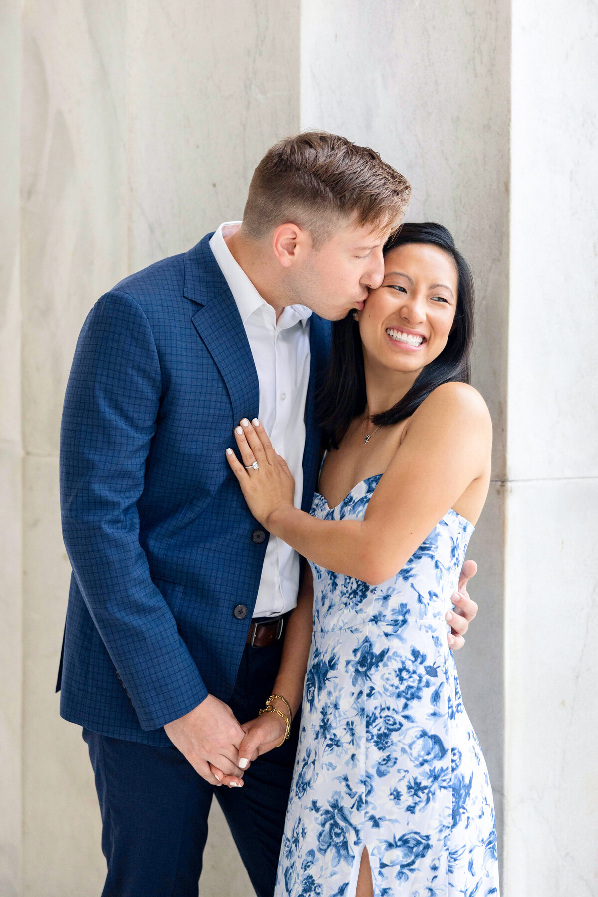 A couple standing against a marble wall, both smiling. The man, wearing a blue suit, is gently kissing the womans temple. The woman, in a white and blue floral dress, has her hand on his chest. They are holding hands, looking happy and in love.
