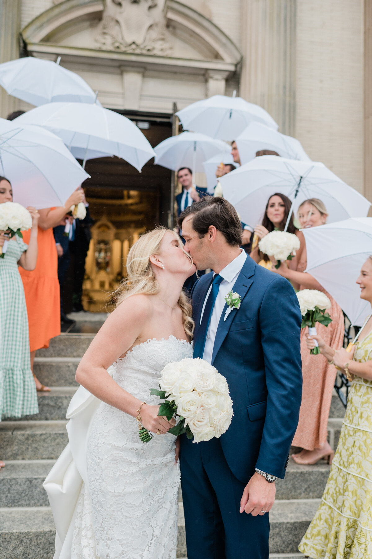 The newlyweds kiss outside Spring Lake Golf Club, with guests in colorful dresses and suits holding white umbrellas and white rose bouquets in celebration