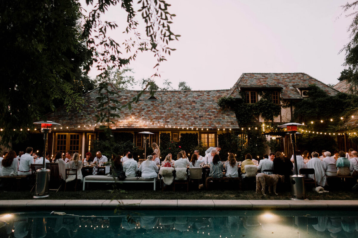A moody and romantic  outdoor alfresco wedding dinner  featuring guests in all white, and pretty twinkly lights hanging overhead.