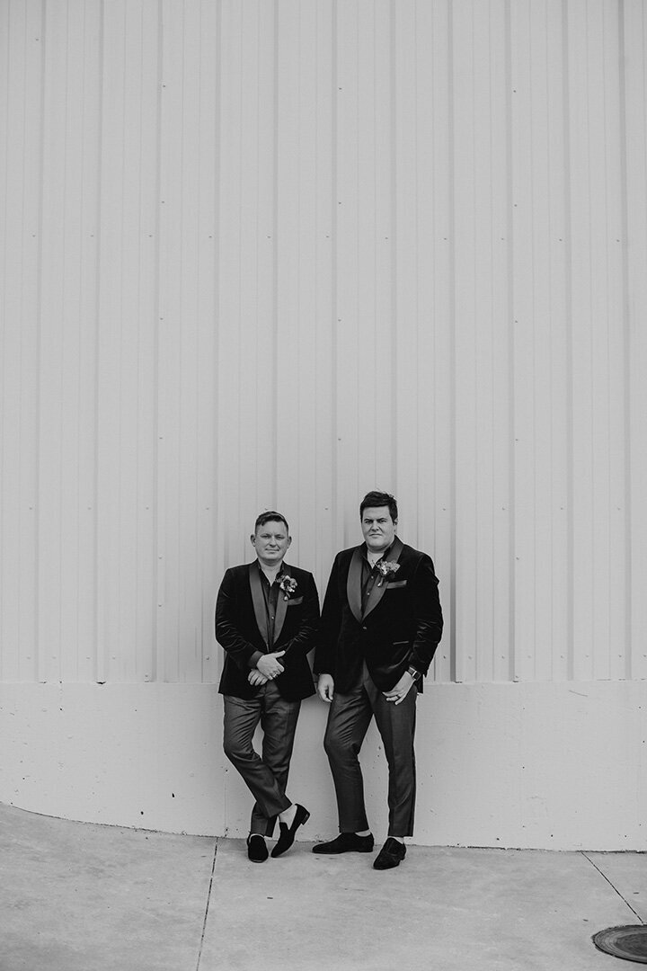 Two grooms wearing tuxedos pose side by side against a white building on a sidewalk.