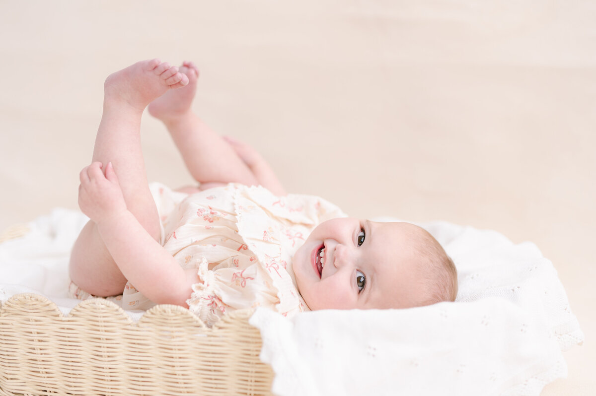 Baby laying in a scalloped basket with a white blanket with her toes up in the air and big smile on her face