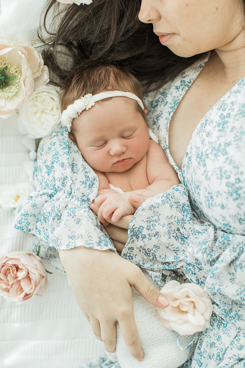 new baby is swaddled laying in bed with asian mom’s arm wrapped around her surrounded by flowers