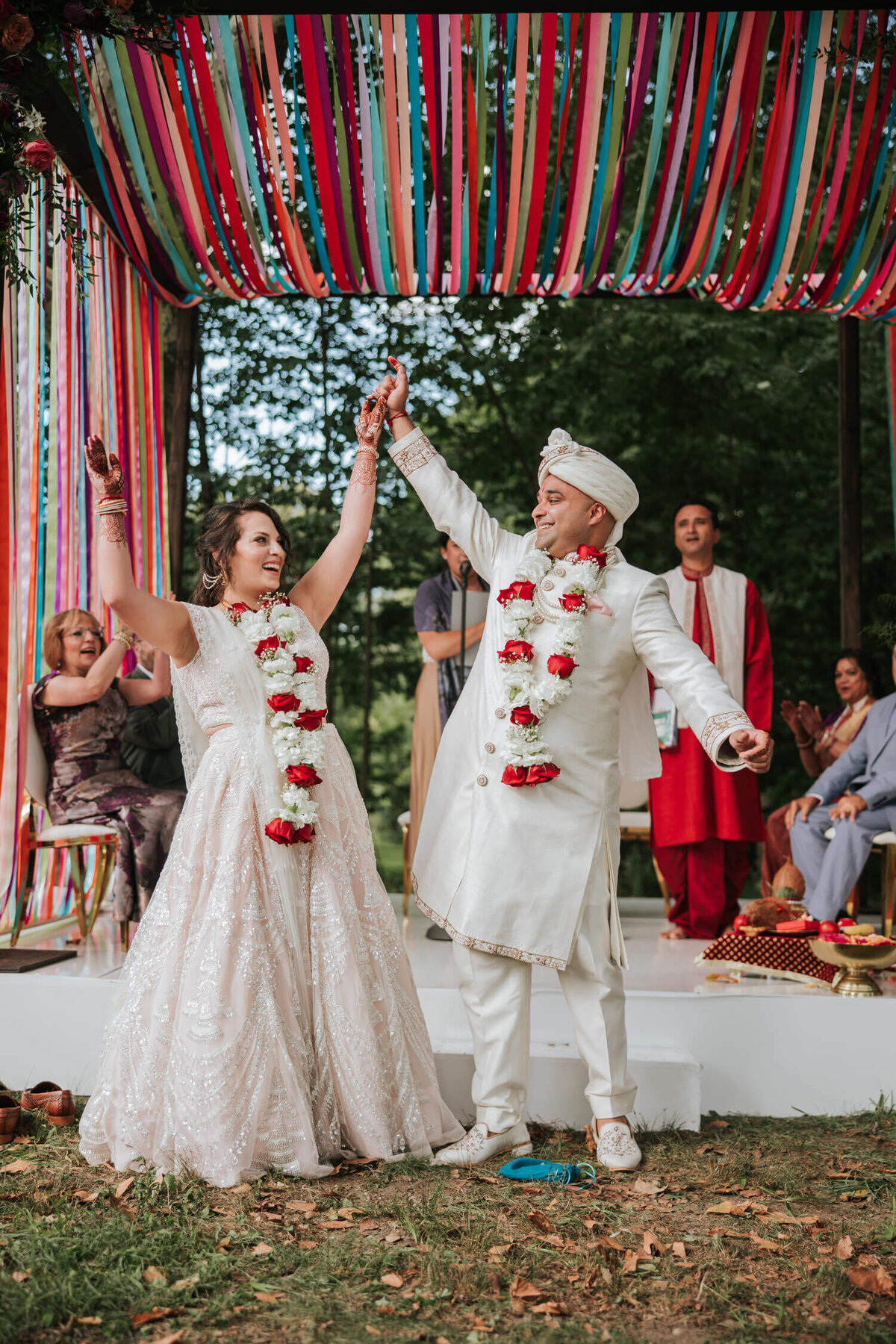 The newlyweds celebrate under a vibrant ribbon canopy, holding hands and smiling radiantly during their joyful outdoor ceremony.