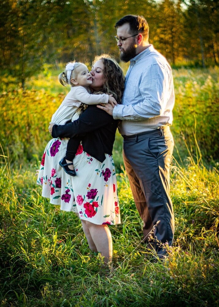 a pregnant woman holds her toddler daughter and gives her a kiss and the dad stands nearby