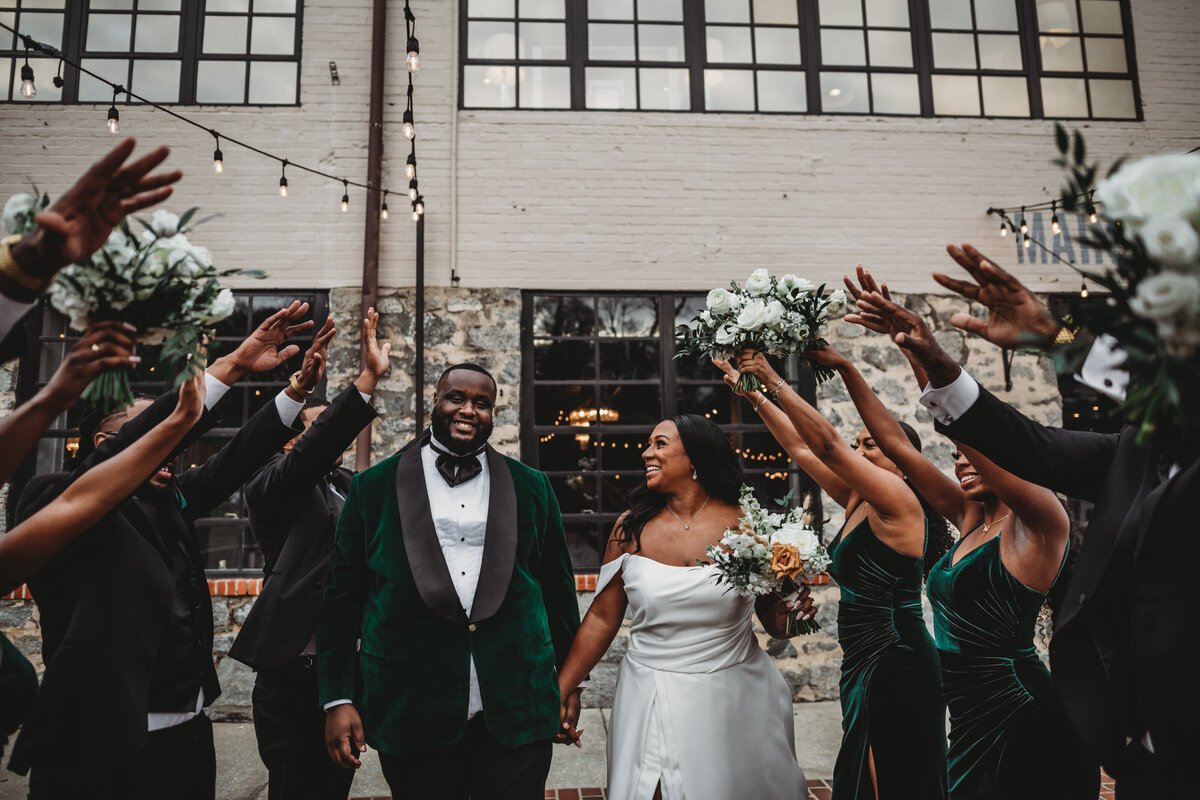 Bride in a satin off the shoulder wedding dress holding a elegant bridal bouquet walks with her groom who is in an emerald green suit as their bridal party holds their hands to create a tunnel for the bride and groom to walk through captured by Baltimore wedding photographers