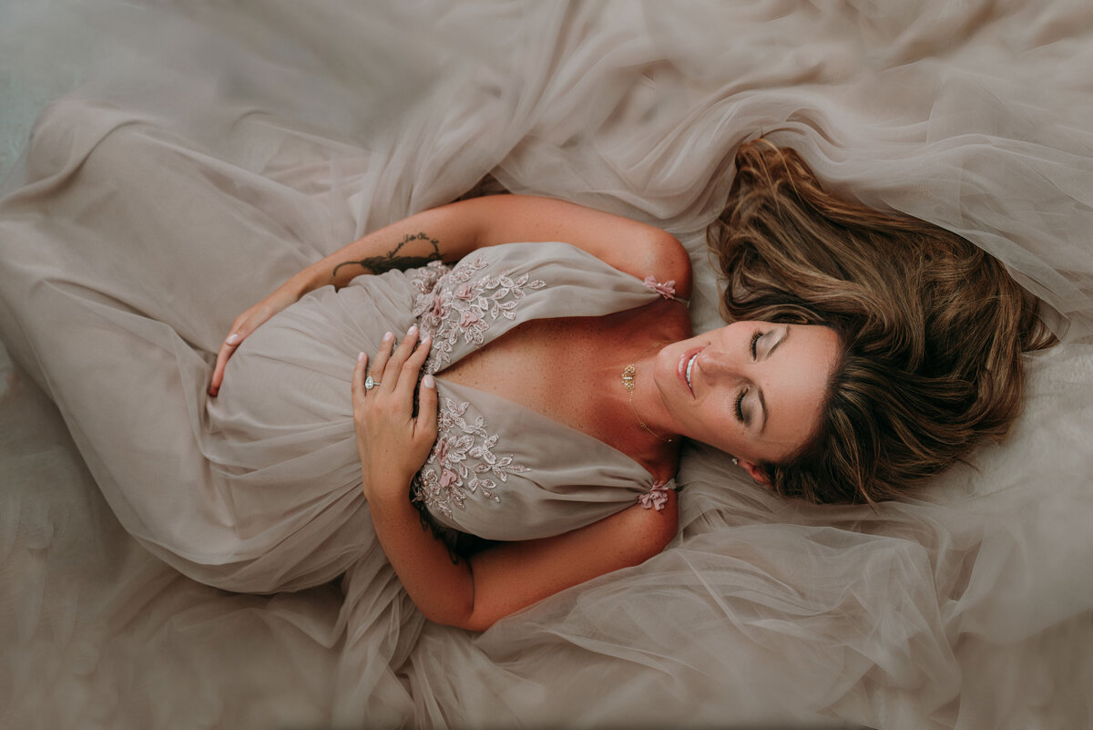 Pregnant woman in gray beaded flowy gown laying down holding baby bump and smilinng
