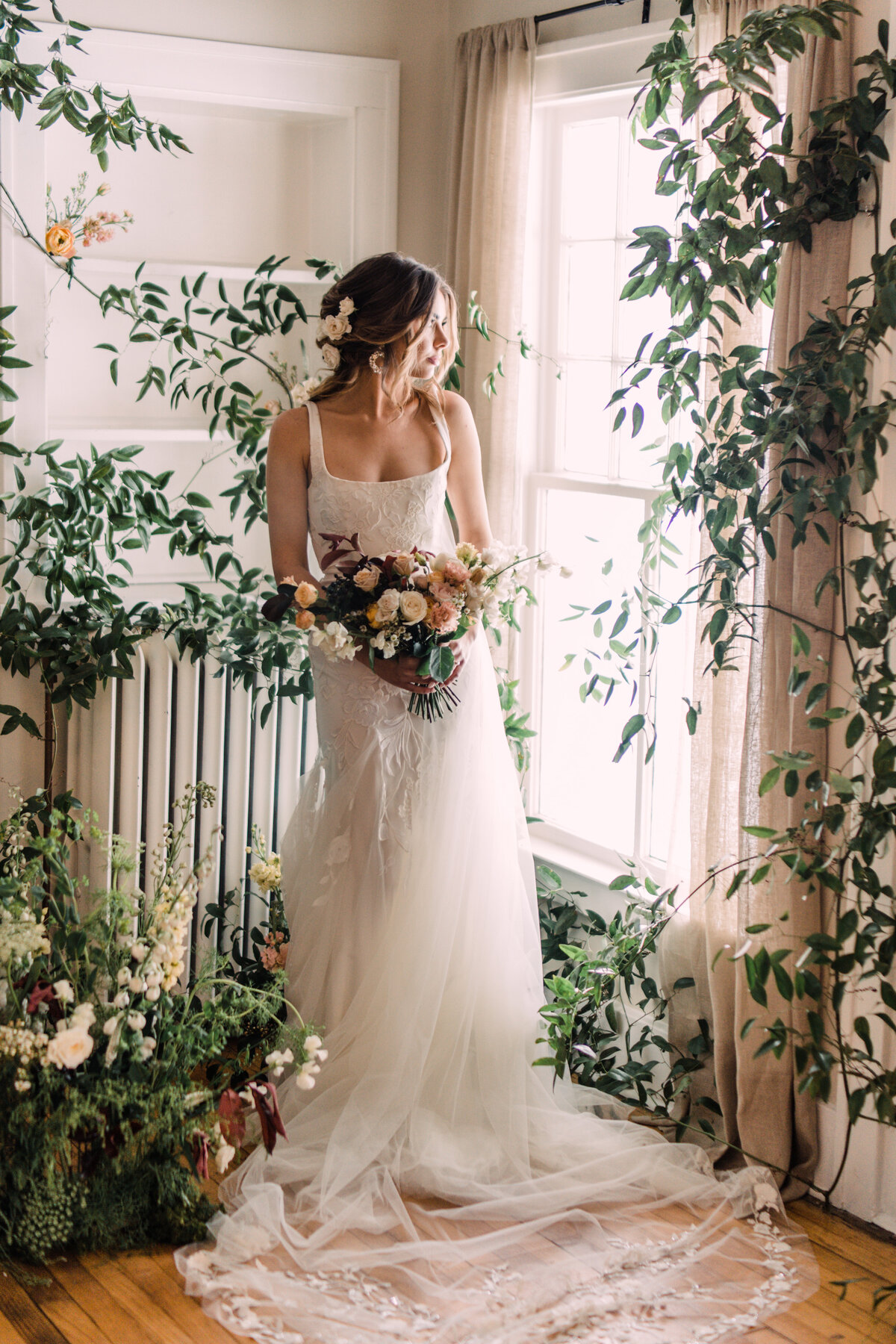 Bride in Alexandra Grecco Marcelle Gown & Ainsley veil holding bouquet in front of window covered in vines at Cornman Farms