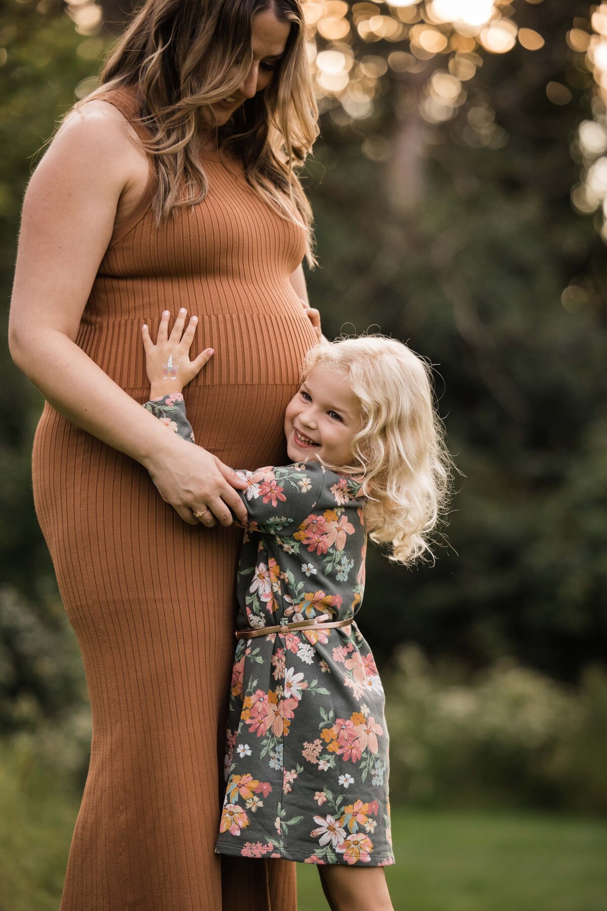 A smiling young child embraces a pregnant woman's belly in an outdoor setting, captured in a stunning piece of maternity photography.