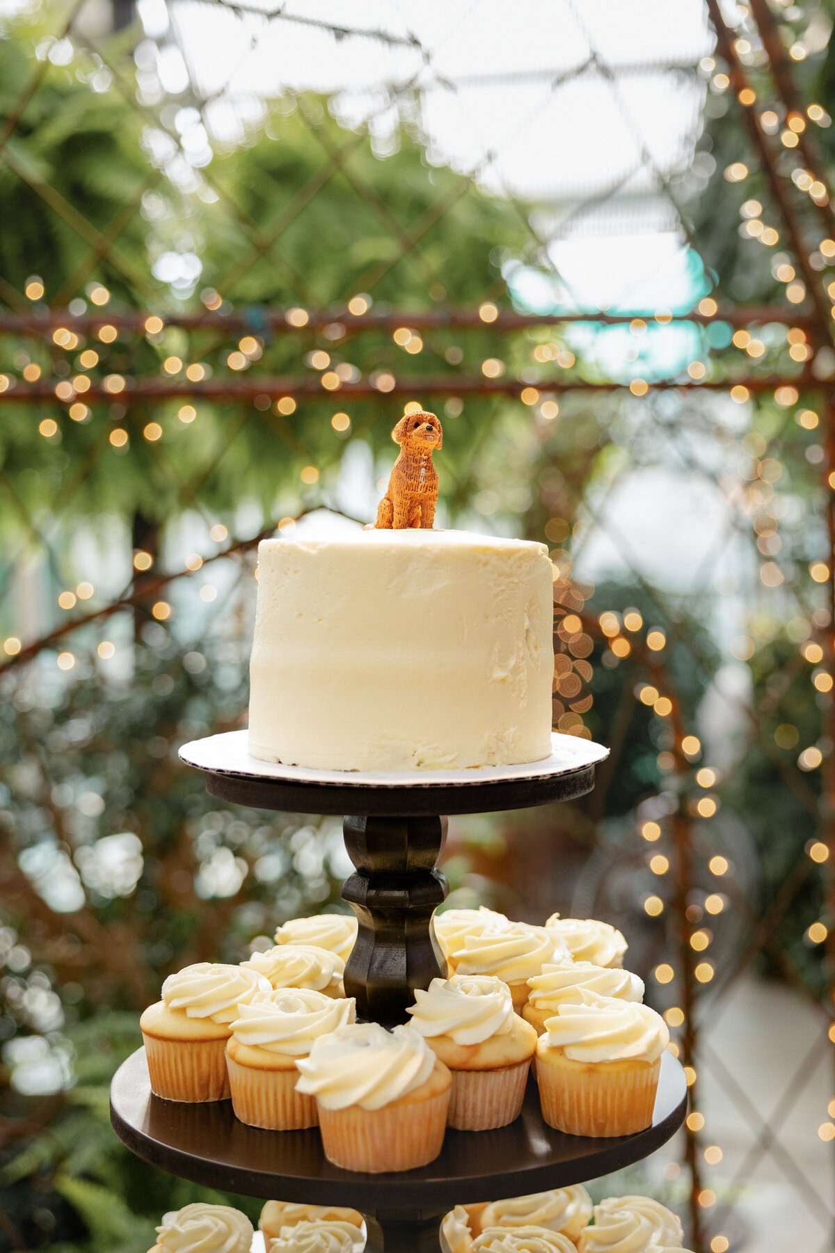 A single-tier wedding cake with a minimalist white design, topped with a small dog figurine, displayed on a dessert table with cupcakes.