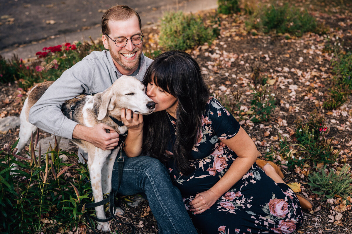 A couple sitting outdoors in a garden, both smiling happily as they cuddle their beagle. The woman is kissing the dog's head while the man holds it close.