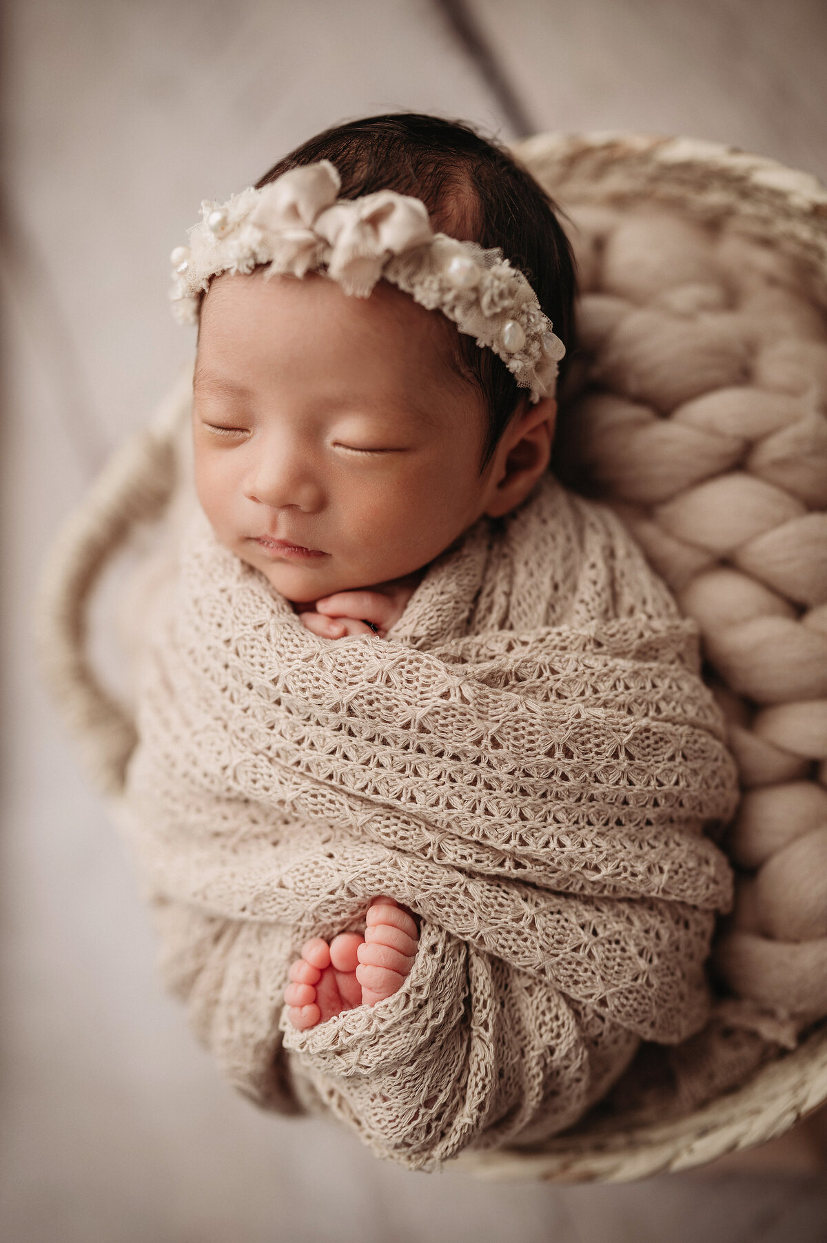 sleeping newborn baby girl wrapped in  textured cream swaddle with feet poking out posed inside of  white wicker basket over white wooden floor
