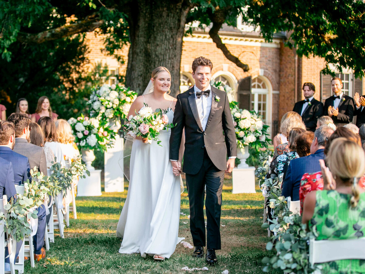 A bride and groom, dressed in a white gown and a black tuxedo, walk hand in hand down an outdoor aisle lined with guests. They are smiling, surrounded by greenery and floral arrangements, with a large tree and brick building in the background.