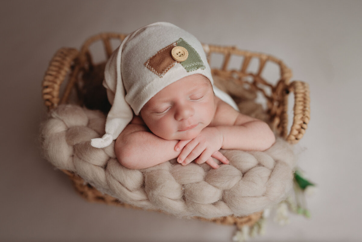 Newborn baby boy propped in brown rattan basket with hands under chin wearing a white sleepy cap