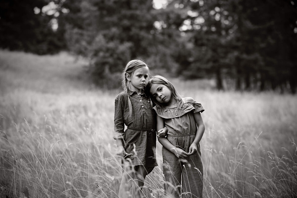 Two sisters stand side by side in a field with one of them resting their head on the others shoulder while looking away longingly. Captured in black and white by Brittany Danielle Photography