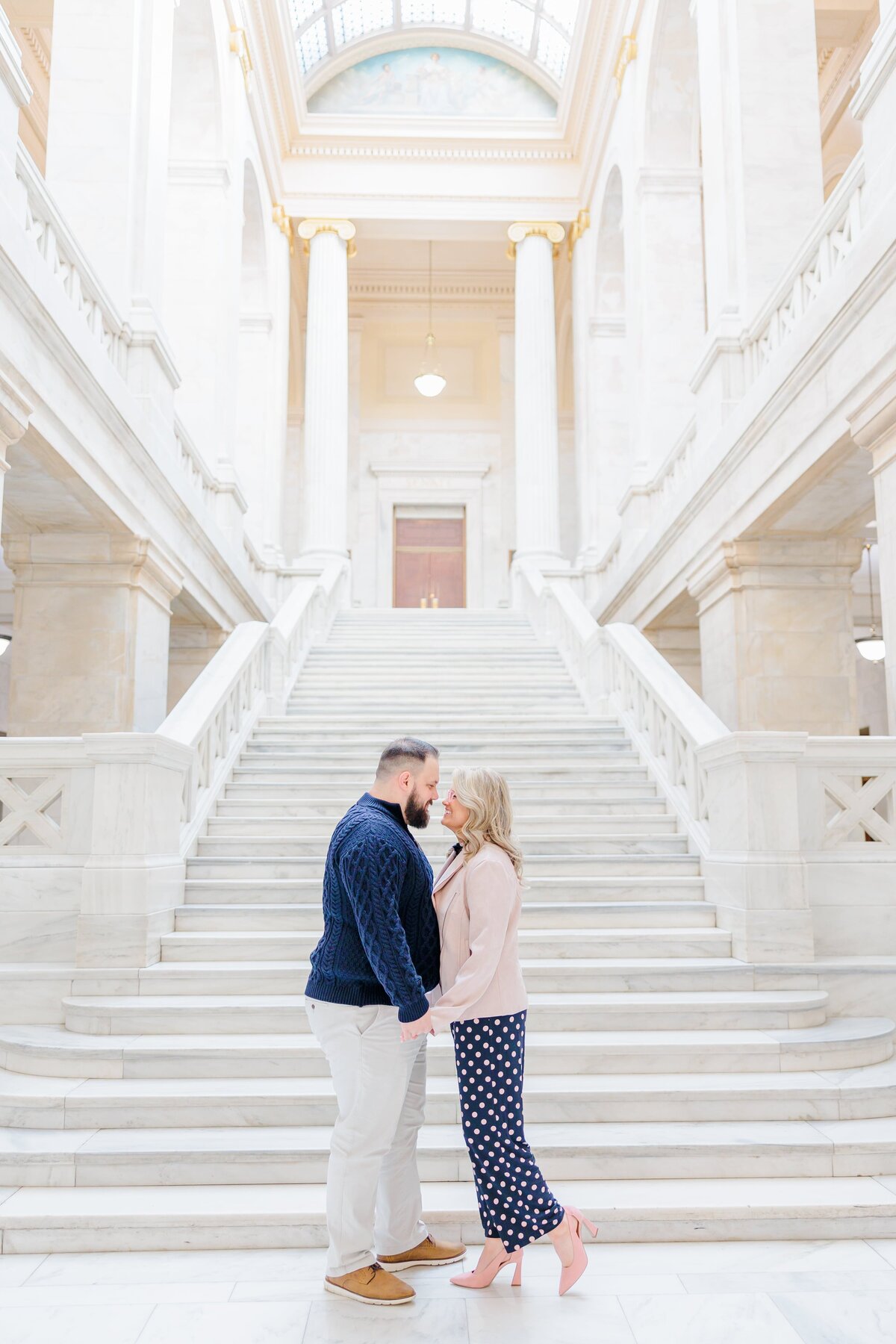 A couple in blue and pink stand inside on the marble steps of the capitol building in Arkansas.