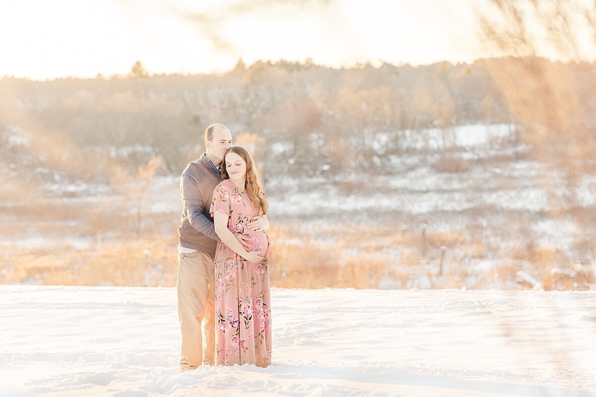 couple stands in the snow during snowy winter maternity photo session with Sara Sniderman Photography in Medfield Massachusetts