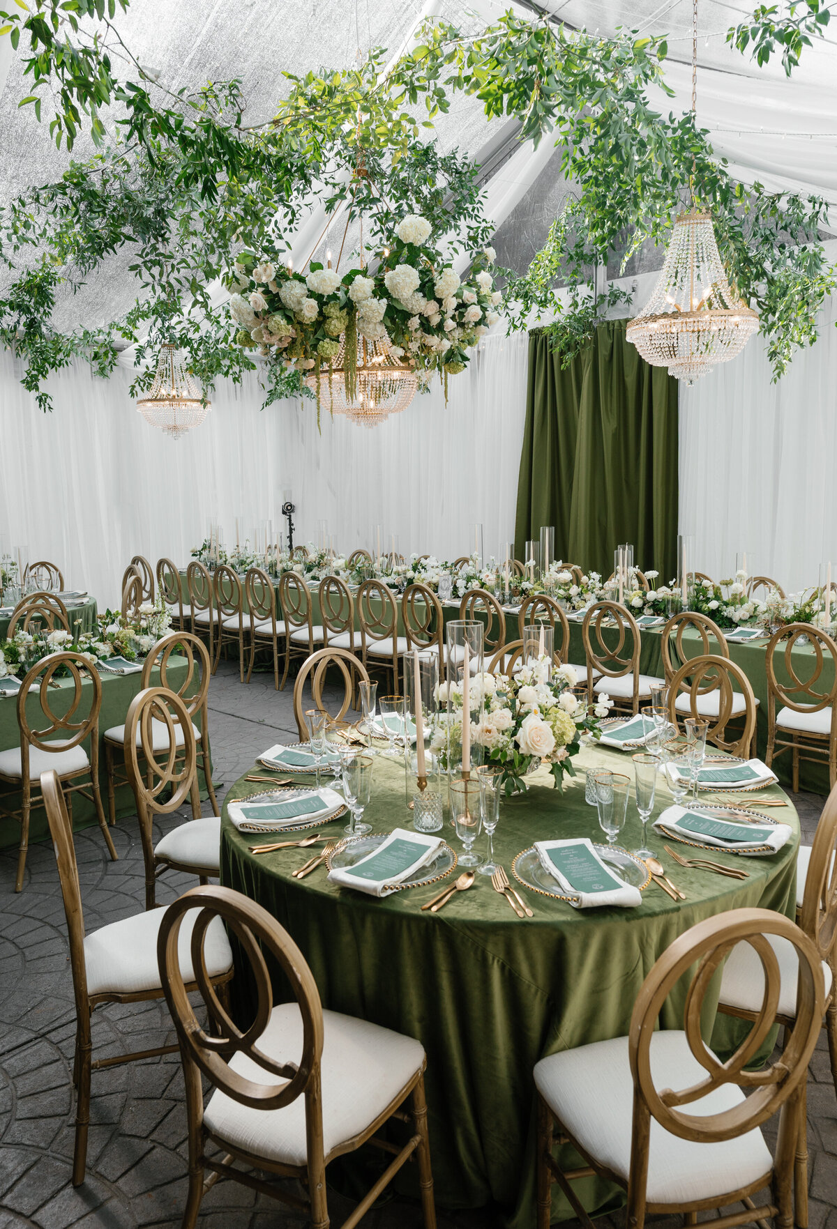 Tented wedding reception featuring round tables with green linens, lush white florals, and elegant crystal chandeliers.
