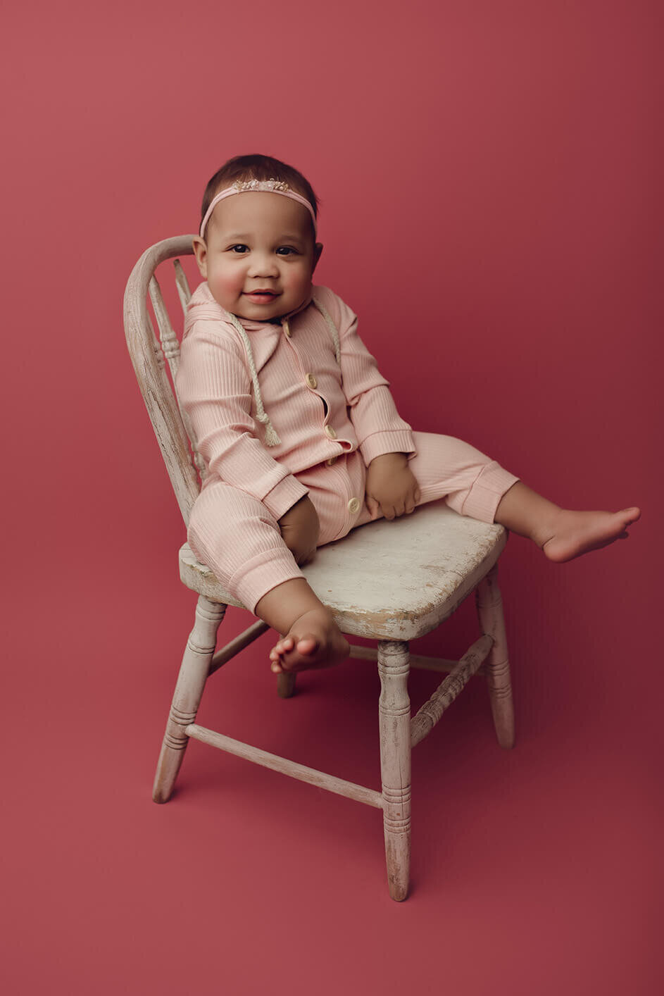 a one year old girl sitting on a white chair in a pink outfit on a pink drop
