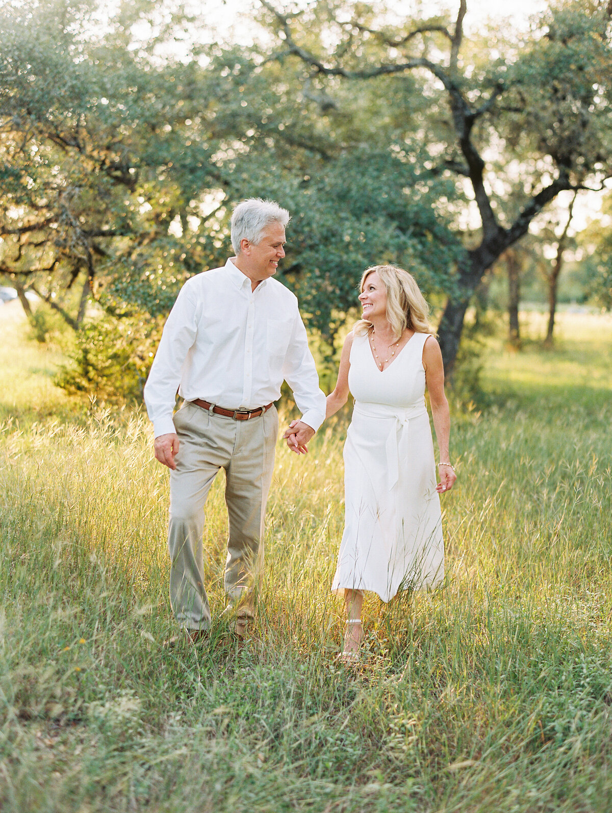 Older couple in white clothes holding hands and smiling at each other while walking through a grassy field