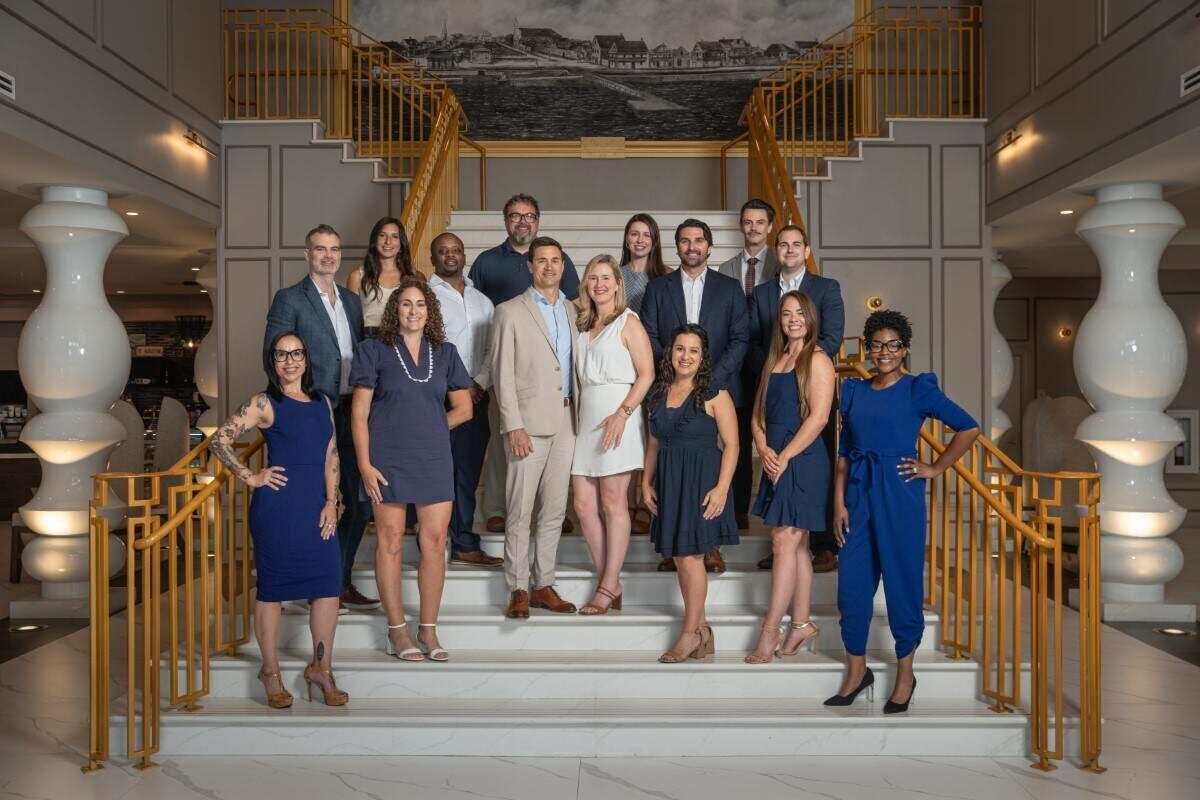 Professional group portrait by St. Augustine headshot photographer featuring nine people in coordinated blue and white attire posed on an elegant curved staircase with ornate railings and classical architecture