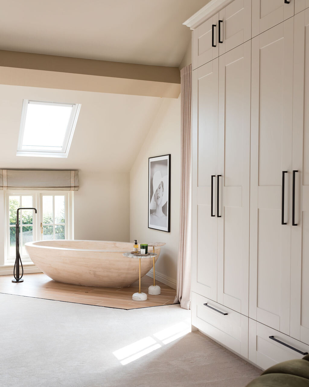 A serene bedroom with a freestanding wooden bathtub on a wooden platform beneath a slanted ceiling with a skylight. There's a window with sheer curtains to the right, a framed black-and-white portrait on the wall, and minimalist decor throughout.