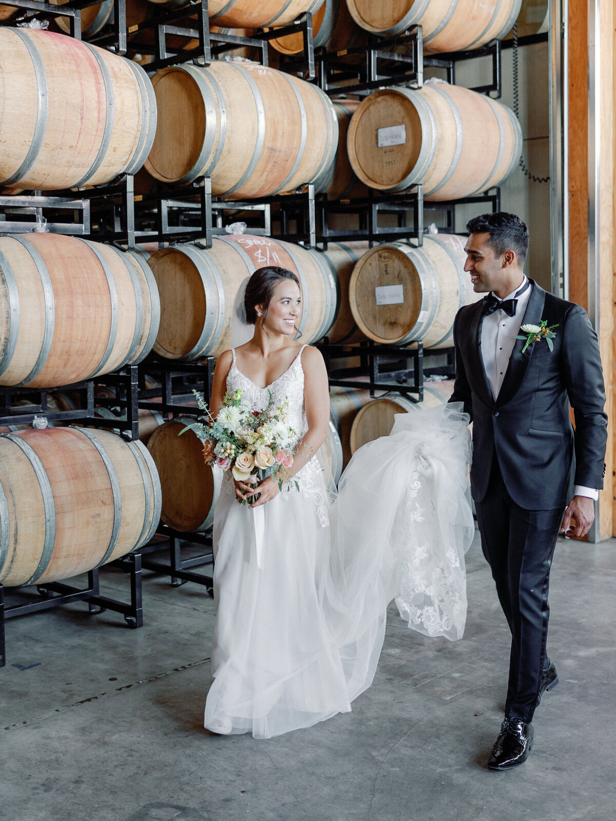 A bride and groom walk together in the wine cellar of district winery while the groom holds his bride's dress train and she looks back at him lovingly