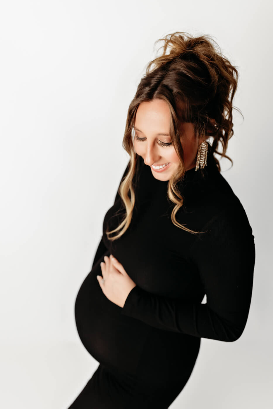 A pregnant woman in a fitted black dress smiles and looks down, touching her belly with one hand. She has wavy hair styled up and wears statement earrings. Perfect for maternity portraits or pregnancy photography on a plain white background.