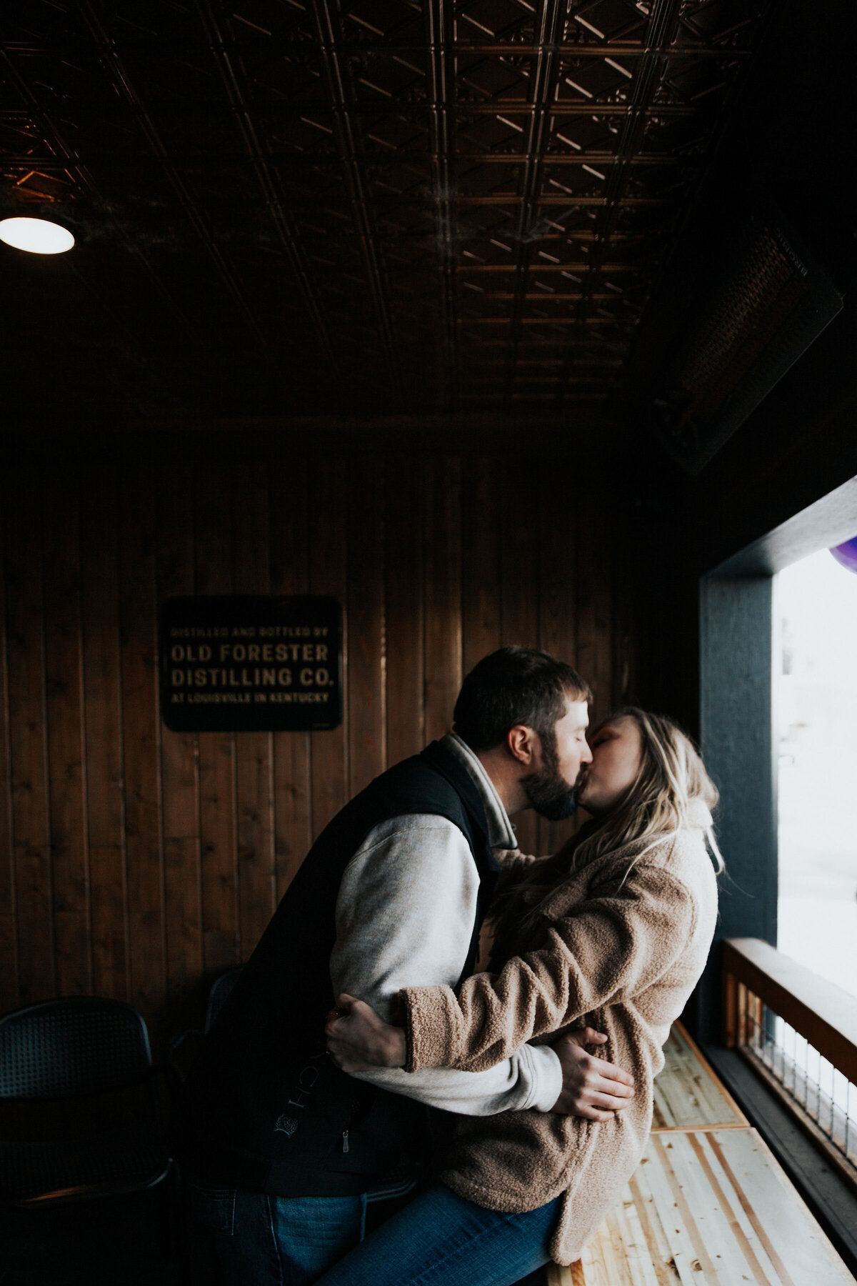 Engaged couple kiss in a patio of a bar while the woman sits on a table top captured by Idaho Falls wedding photographer