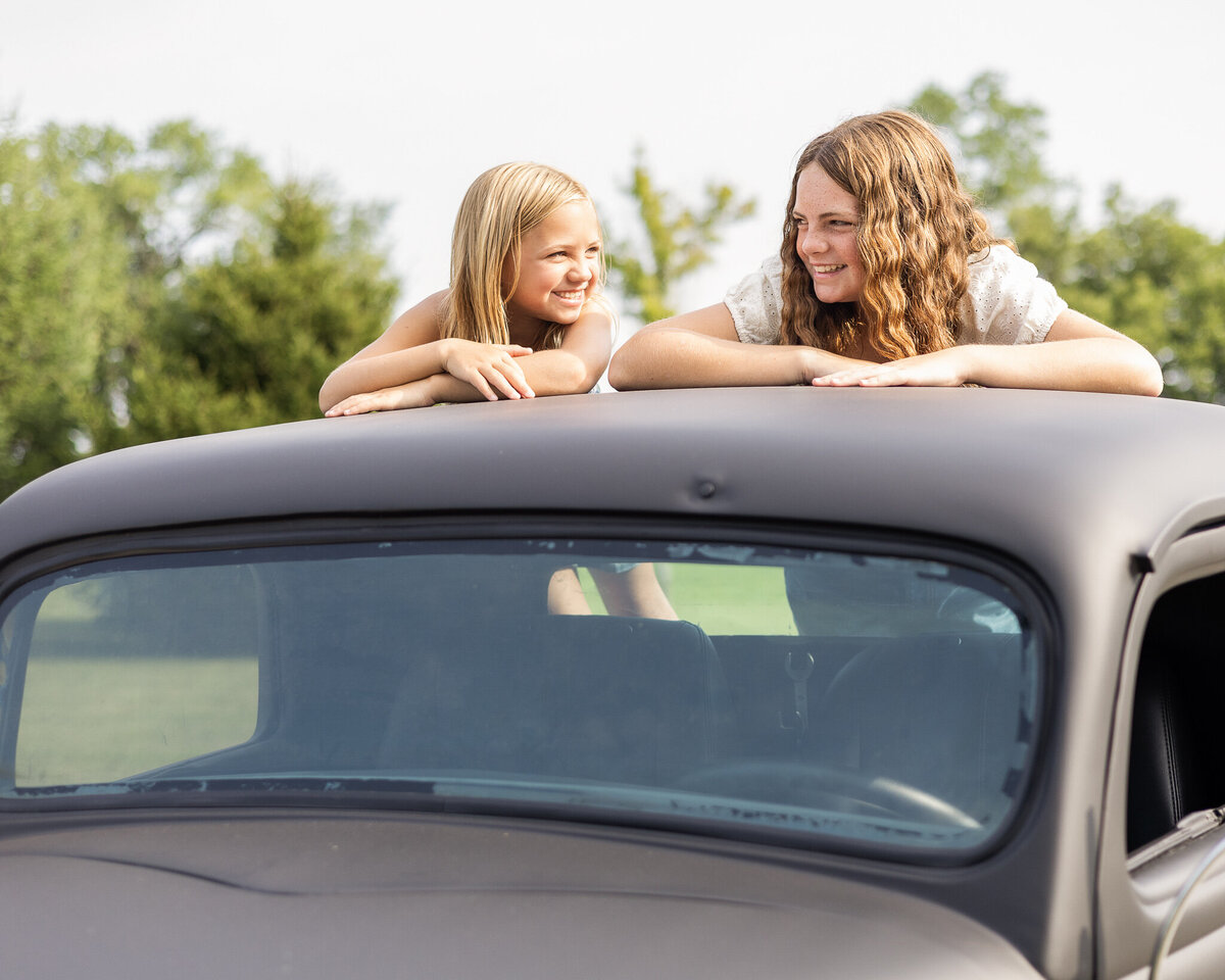 Sisters standing in the bed of the classic black car with their hands on top of the roof, looking at each smiling.  Younger sister has medium length blonde hair and older sister has long brown hair.
