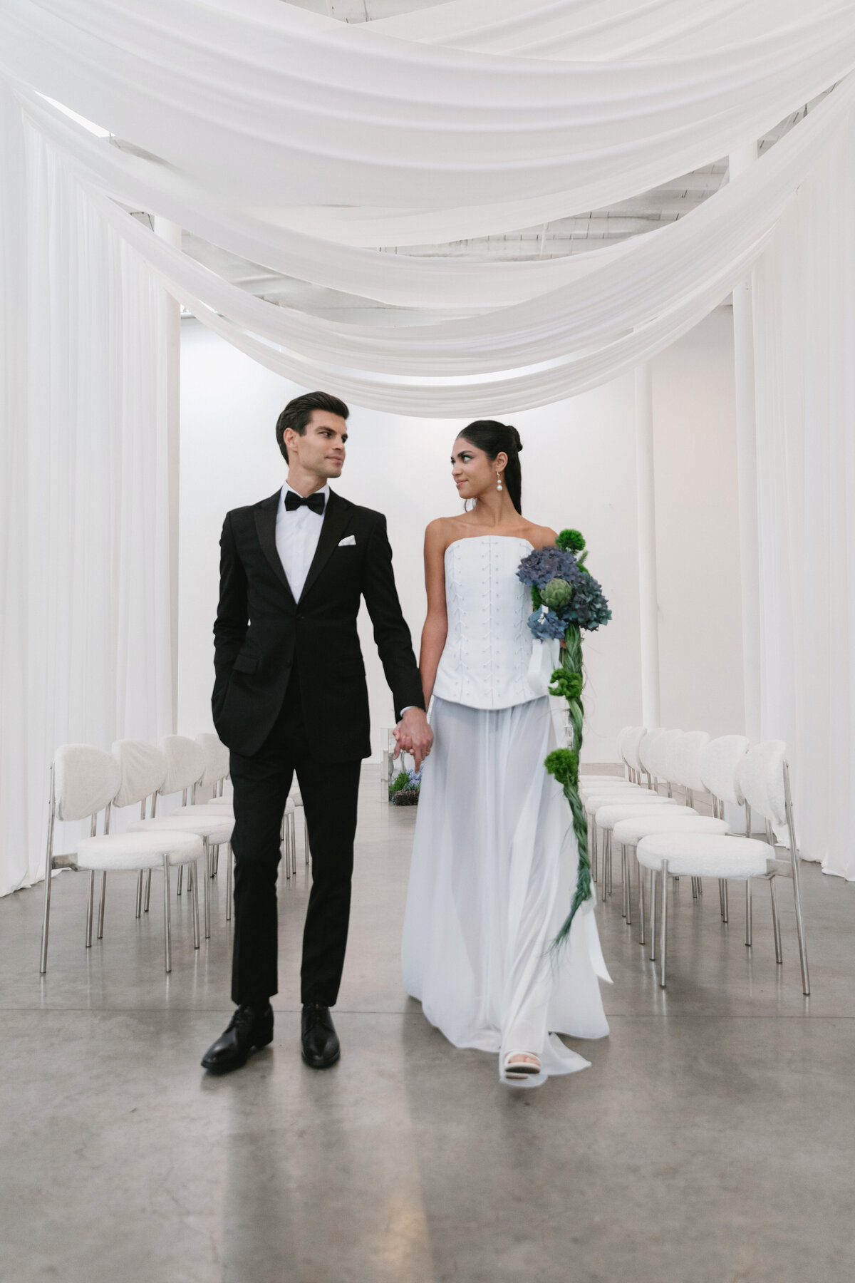 Bride and groom reaching toward each other over a sculptural blue and green floral arrangement during an editorial NYC wedding shoot.