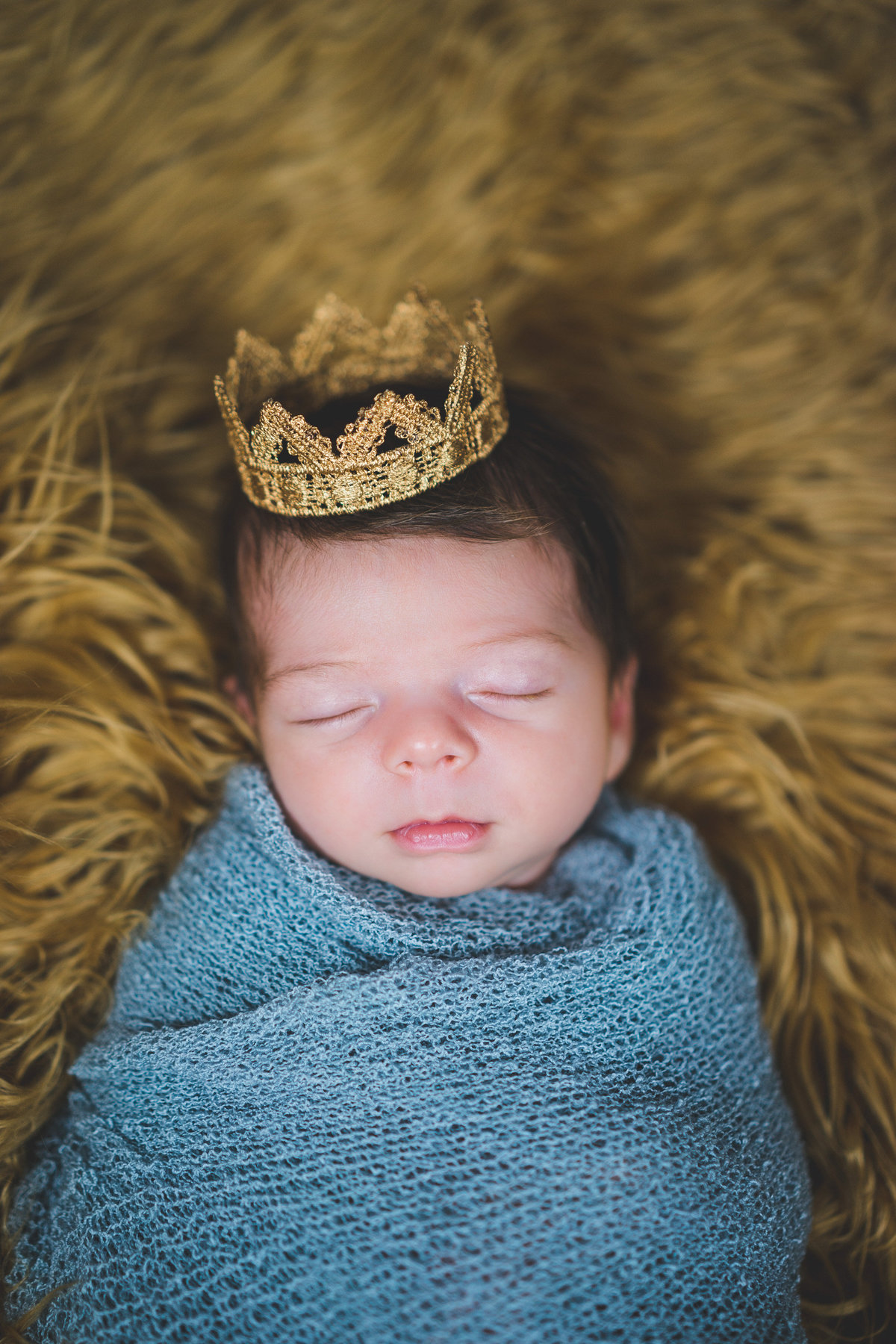 Newborn baby wrapped in cheesecloth blanket wearing a gold crown laying on furry blanket in San Antonio Photography studio.