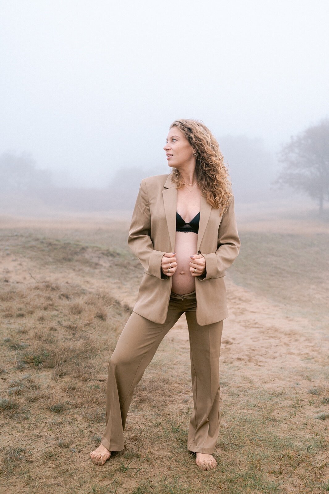 Zwangere vrouw houdt haar blazer beet terwijl ze op zij kijkt. Ze kijkt naar de zijkant.