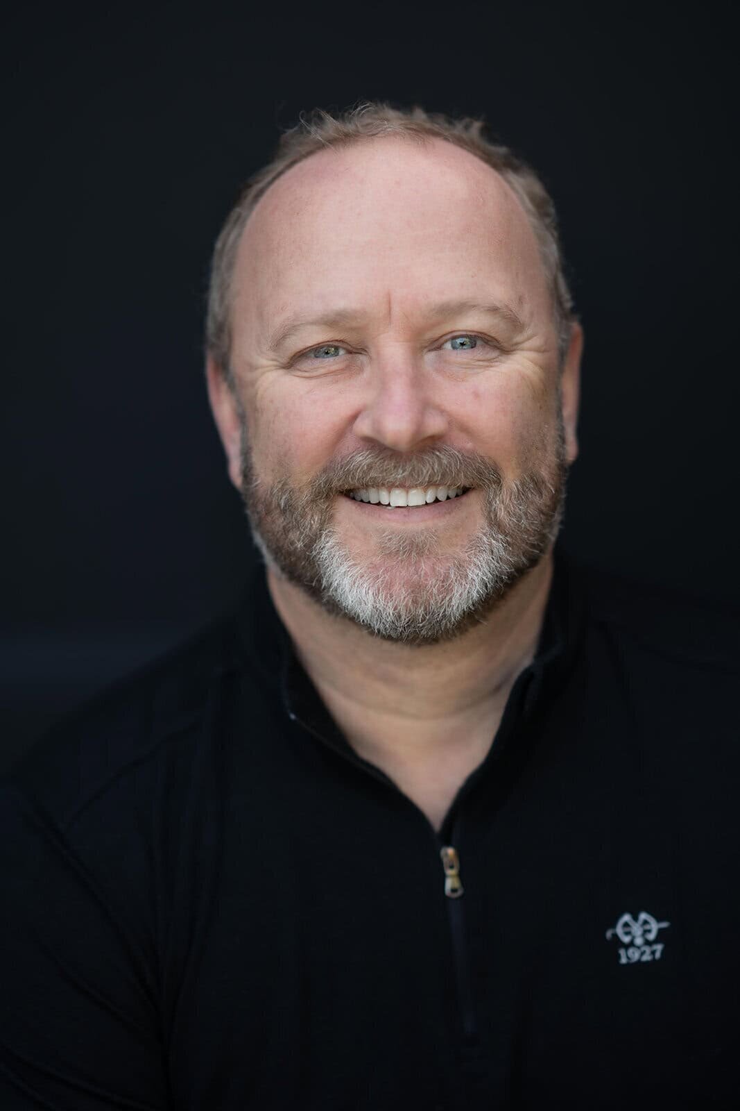 Distinguished looking gentleman with a beard wearing a dark shirt against a dark background