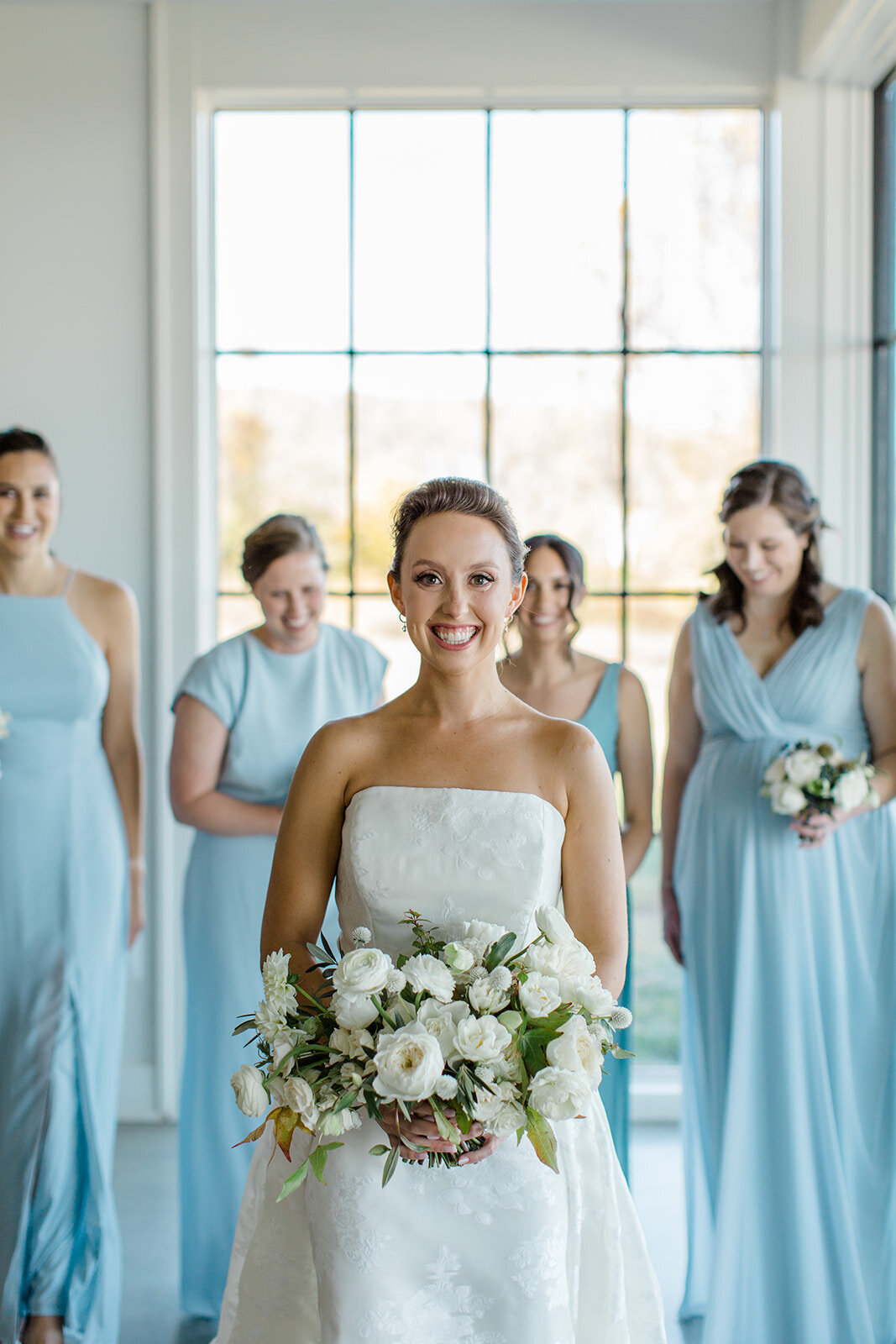 Bride smiling wearing her white gown while smiling with her maids behind wearing blue gown while holding their bouquets