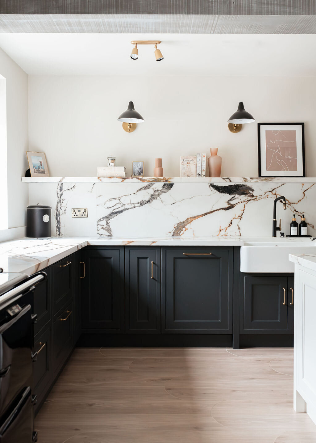 A modern kitchen with dark lower cabinets, a white marble backsplash with black and gold veining, a farmhouse sink, and minimalist open shelving holding books and decor. Two wall-mounted lights illuminate the space, which also features light wood flooring.