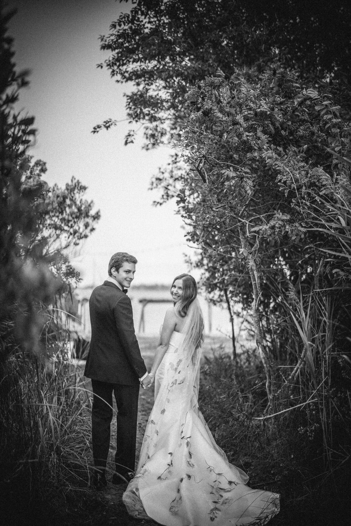 A couple in formal attire holds hands and looks back while walking on a path surrounded by trees and foliage. This photo was taken by a destination wedding photographer for Tuscan vineyard weddings