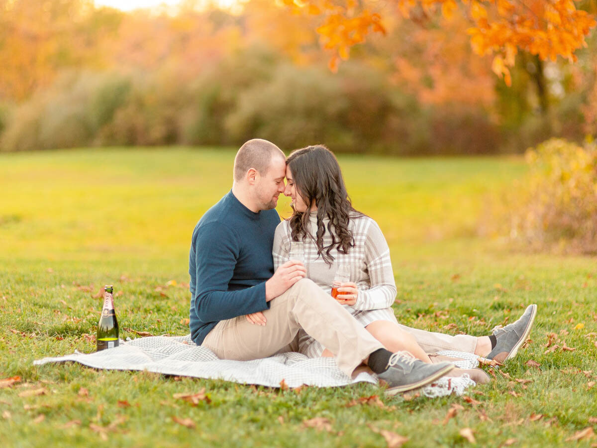 Man and woman drink champagne as they laugh during their picnic engagement session.
