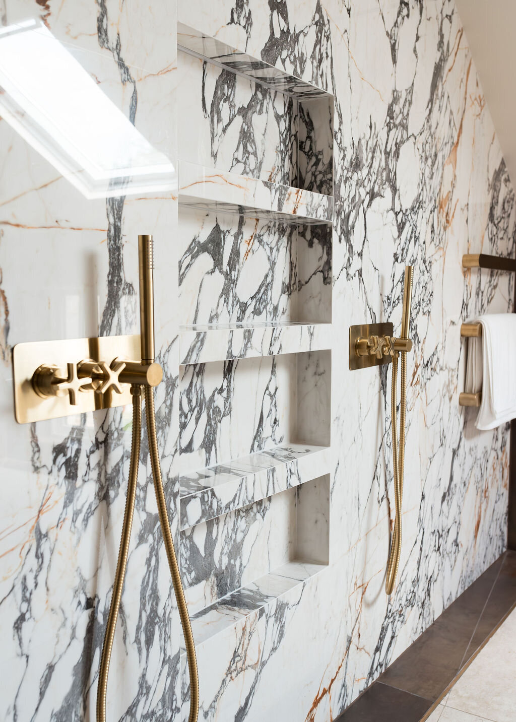 Two modern gold shower fixtures mounted on a white marble wall with black veining. Between them, multiple shelves are built into the marble wall. To their right are three gold towel bars.