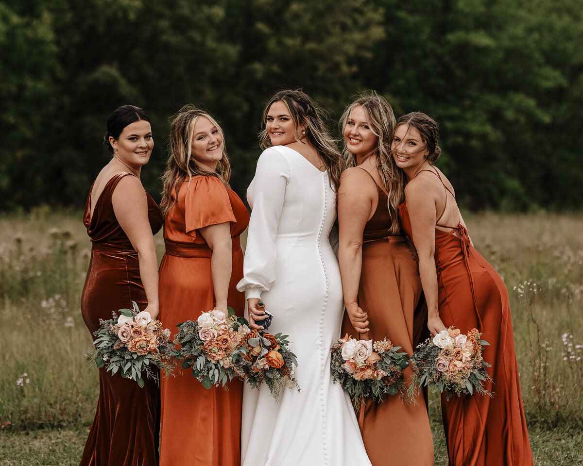 A radiant bride in white with her bridesmaids in coordinated terracotta dresses, holding bouquets, sharing a joyful moment outdoors taken by jen Jarmuzek photography a Minneapolis wedding photographer