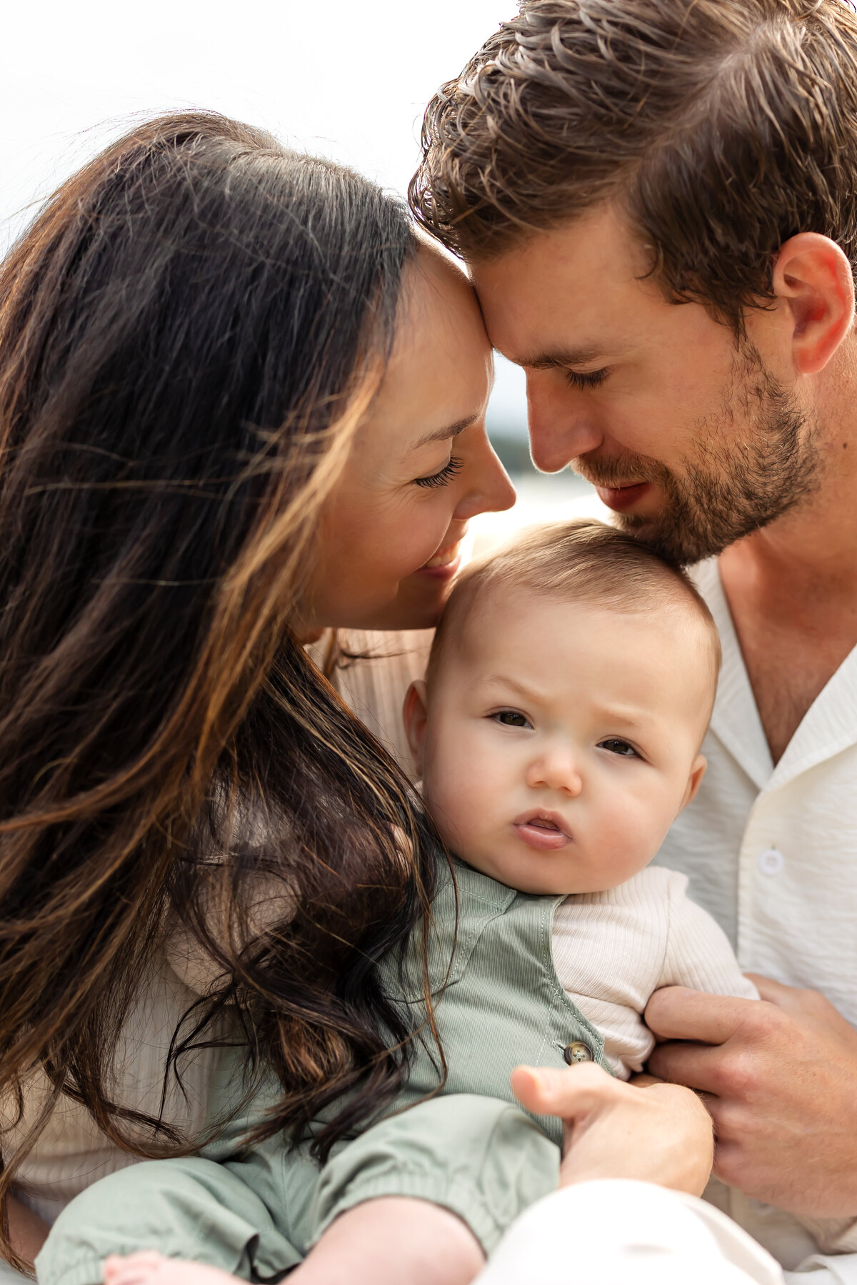a mother and father touch noses to eachother as they smile down at their baby who is smiling at the camera