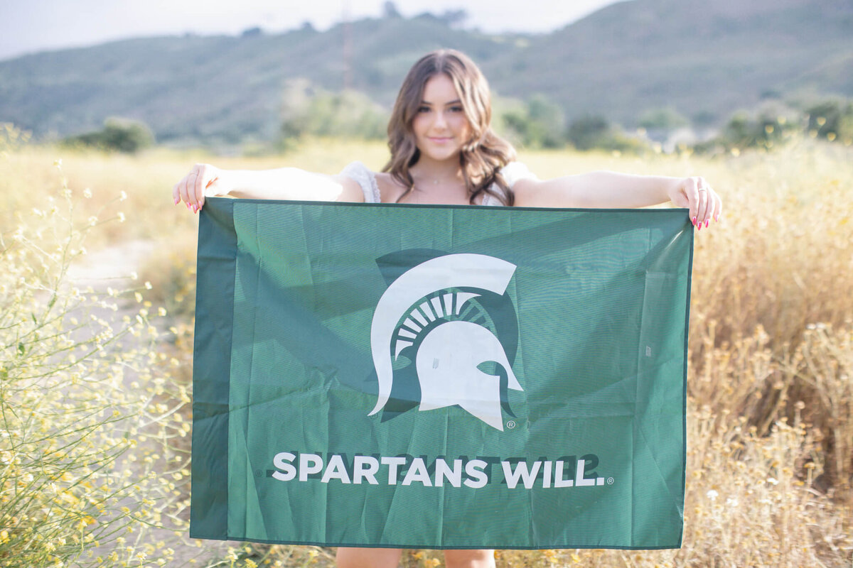 A college graduate with long, brown hair holding up a green flag with the Michigan State logo on it and the words "Spartans Will," looking at the camera and standing in a field of grass in San Juan Capistrano.