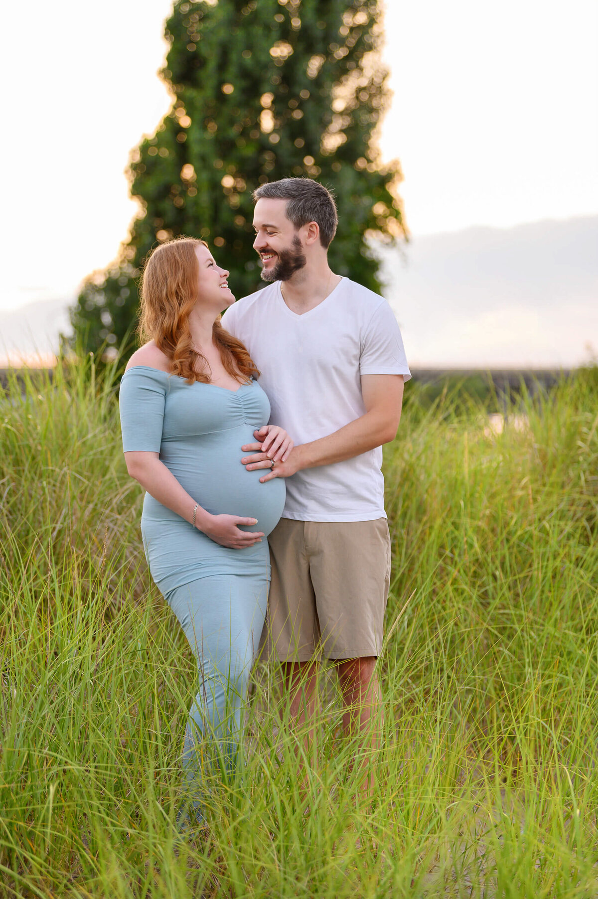 husband and wife looking at each other while holding mom's belly at their outdoor beach maternity photography session in Burlington, On