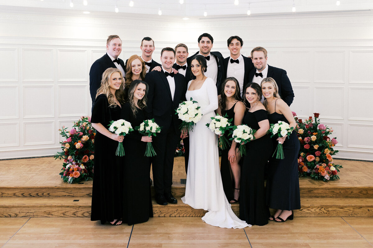 Wedding party group in elegant black attire, posing with smiles at St. Paul College Club. The bridesmaids hold beautiful bouquets of roses, and the groomsmen wear bowties, all surrounded by elegant florals. Captured by Toly Dzyuba Photography, Minnesota wedding photographer.
