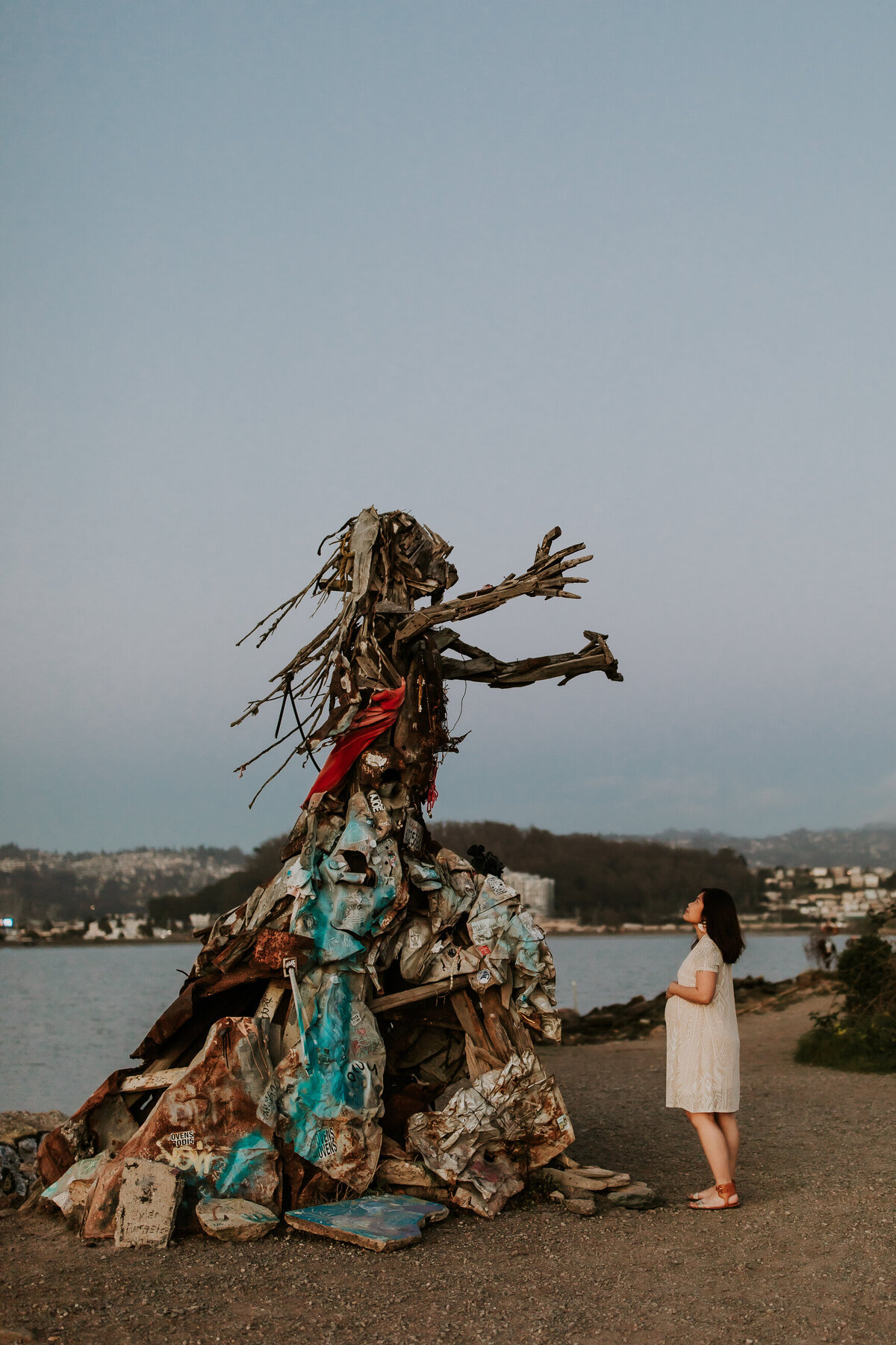 Bay Area pregnant mom in white dress looks up at large art sculpture of women with open arms at albany bulb